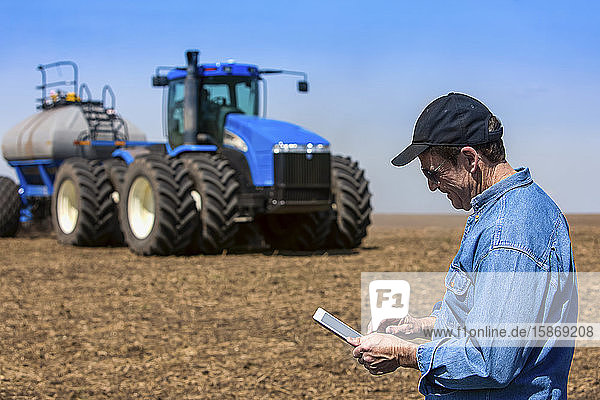 Farmer using a tablet while standing on a farm field and a tractor and equipment seeds the field; Alberta  Canada