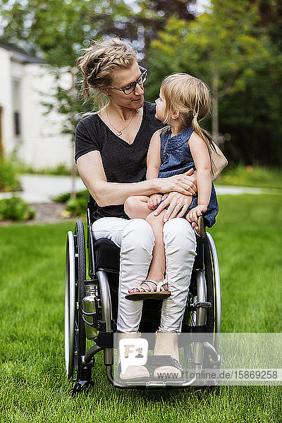 A paraplegic mom holding her little girl in her lap while sitting in her wheelchair in her front yard on a warm summer afternoon: Edmonton  Alberta  Canada.