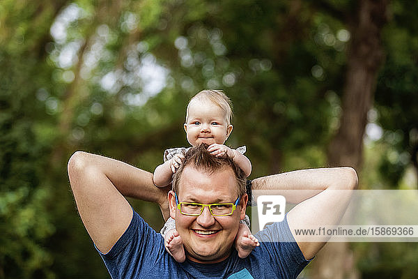 A father holding his baby girl on his shoulders and she is holding his head  while outdoors during the fall; Edmonton  Alberta  Canada