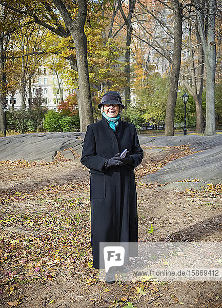 Ältere Frau steht für ein Porträt im Central Park im Herbst  hält ein Smartphone und trägt einen langen Mantel; New York City  New York  Vereinigte Staaten von Amerika