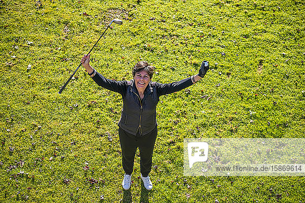 Hohe Winkelansicht einer Golferin auf grünem Gras  die ihren Schläger und Ball in die Luft hält und zur Kamera aufblickt; Schweiz