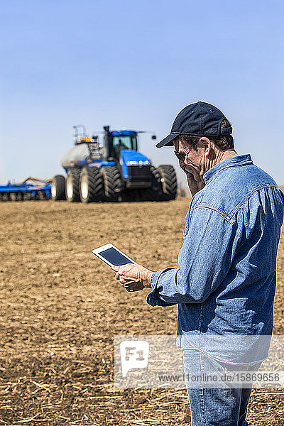 Farmer using a smart phone and tablet while standing on a farm field and watching the tractor and equipment seeding the field; Alberta Canada
