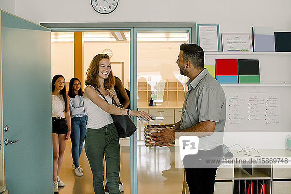 Smiling teenage girl giving phone to teacher while walking in classroom