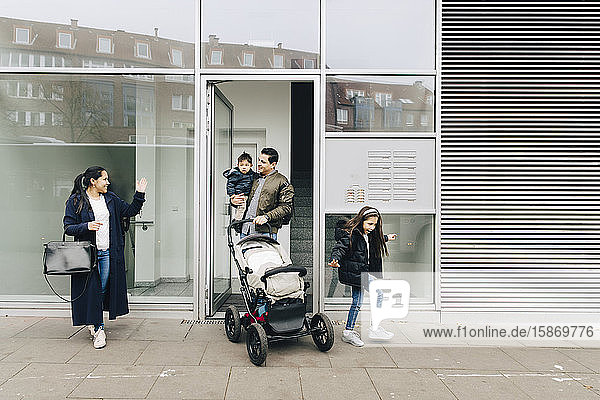 Mutter winkt Vater und Kindern mit Kinderwagen zu  während sie in der Stadt auf dem Bürgersteig geht