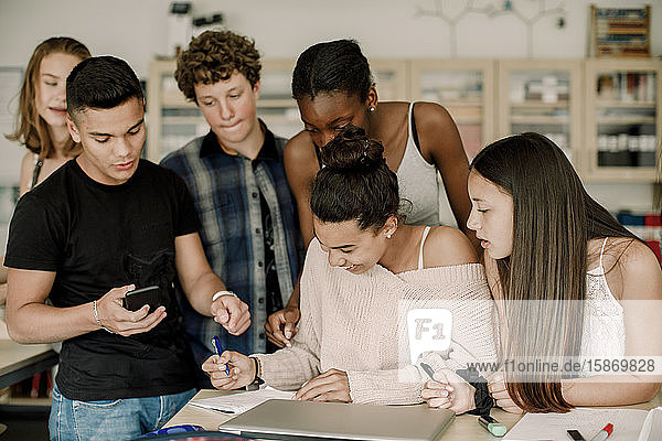 Smiling teenager studying while friends standing by table in classroom
