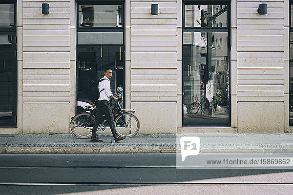 Geschäftsmann in voller Länge zu Fuß mit dem Fahrrad auf dem Bürgersteig gegen Gebäude in der Stadt