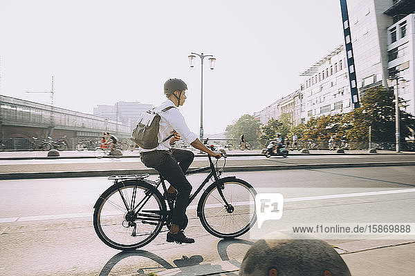 Männlicher leitender Angestellter mit Rucksack fährt Fahrrad auf Straße in Stadt gegen Himmel