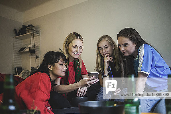 Lächelnde Freundinnen  die ein Mobiltelefon benutzen  während sie zu Hause sitzen