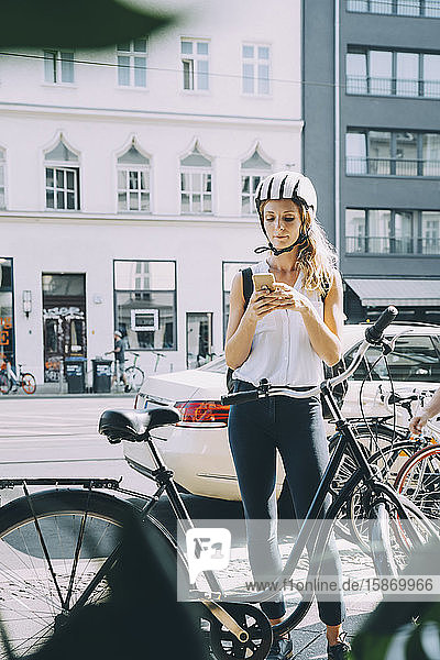 Geschäftsfrau mit Helm  die mit ihrem Mobiltelefon auf der Straße in der Stadt steht