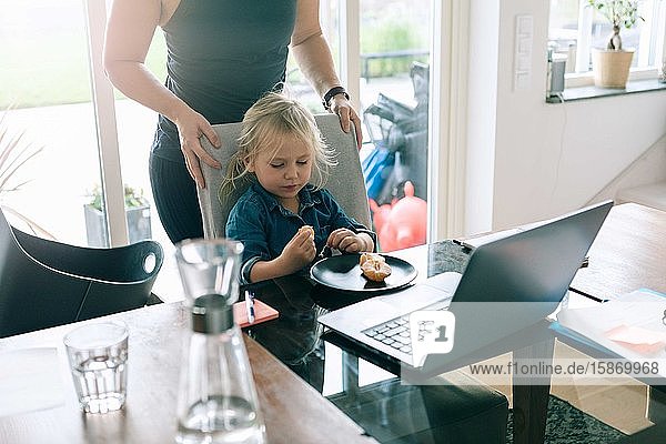 Mädchen isst Obst  während die Mutter zu Hause hinter einem Stuhl steht