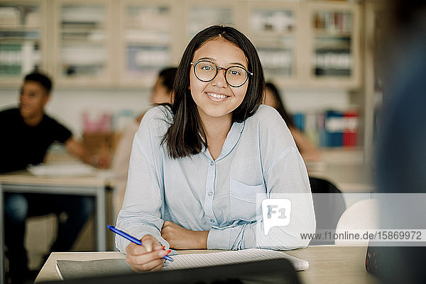 Porträt einer lächelnden Teenagerin mit Brille beim Lernen am Tisch im Klassenzimmer