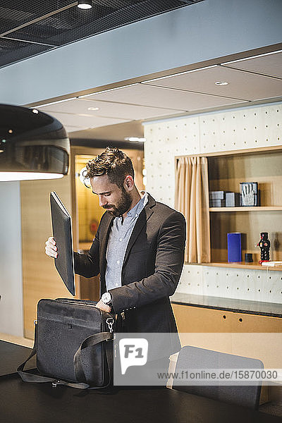 Young businessman keeping laptop in bag while standing by table in office