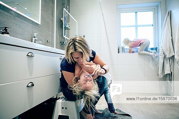 Smiling mother brushing daughter's teeth while girl climbing on window in bathroom