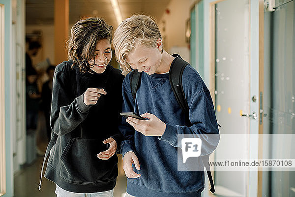 Lächelnde männliche Schüler mit Smartphone auf dem Schulkorridor