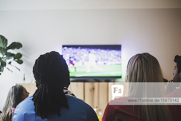 Rückansicht von Freunden  die sich ein Fußballspiel ansehen  während sie im Wohnzimmer sitzen