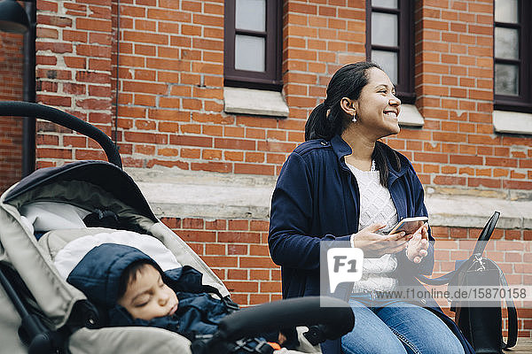 Lächelnde Mutter benutzt Smartphone  während Sohn im Kinderwagen gegen Gebäude in der Stadt sitzt