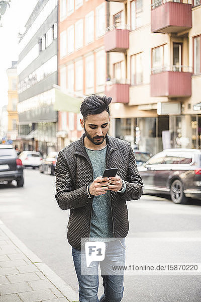 Junger Geschäftsmann benutzt Smartphone beim Gehen auf dem Bürgersteig in der Stadt