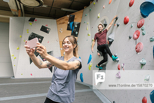 Lächelnde Frau  die sich mit einem männlichen Freund über ein Smartphone selbstständig macht  klettert im Fitnessstudio an einer Wand