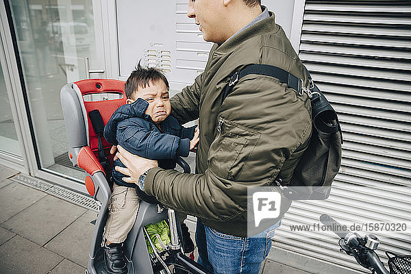 Mittelteil eines Vaters  der einen weinenden Sohn hält  der auf einem Fahrradsattel am Bürgersteig in der Stadt sitzt