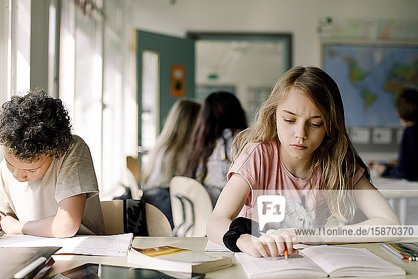 Female student studying from book while sitting by male friend in classroom