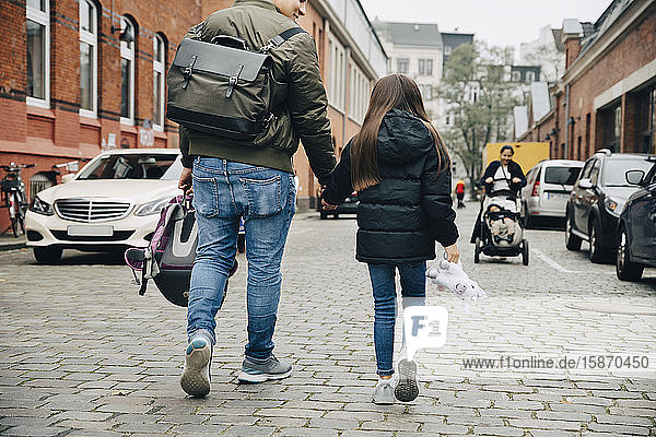 Rückansicht eines Vaters und einer Tochter  die sich bei einem Spaziergang auf einem Fußweg in der Stadt an den Händen halten
