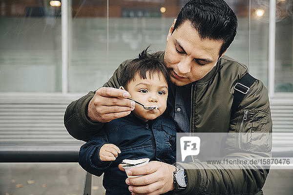 Father feeding baby food to son while sitting on bench in city