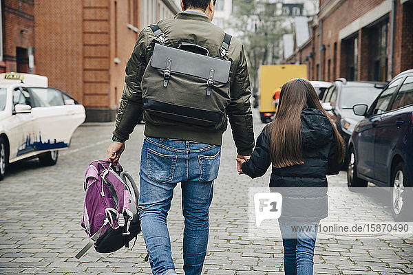 Rear view of father and daughter with backpack walking on footpath in city