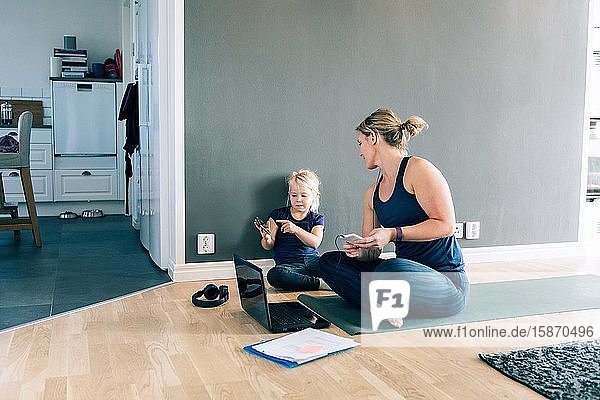 Tochter zeigt mit dem Smartphone auf die Mutter  während sie zu Hause auf der Sportmatte sitzt