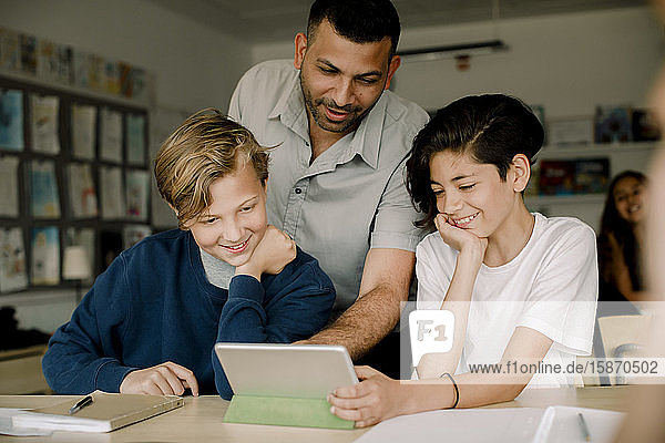 Lächelnder männlicher Tutor mit Schülern  die ein digitales Tablet benutzen  während sie im Klassenzimmer sitzen