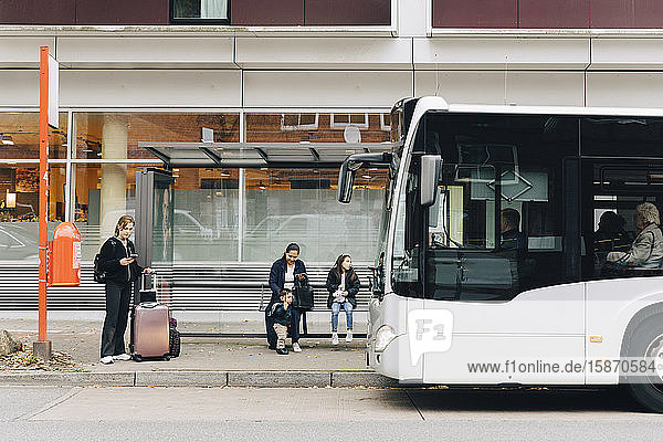 Frau in voller Länge mit Gepäck und Familie wartet an der Bushaltestelle in der Stadt auf den Bus
