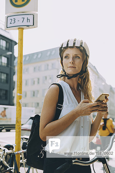 Junge Frau auf dem Fahrrad schaut weg  während sie in der Stadt ein Smartphone in der Hand hält
