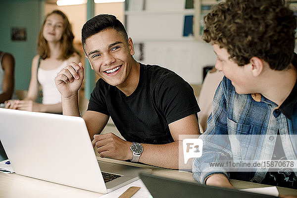 Lächelnde männliche Studenten  die den Laptop über dem Tisch benutzen  während sie im Klassenzimmer sitzen