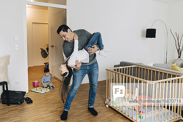 Verspielter Vater in voller Länge  der seine Tochter hochhebt  während der Sohn im Wohnzimmer mit Spielzeug spielt