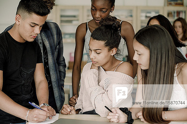 Teenage boy solving while friends looking in book by table in classroom