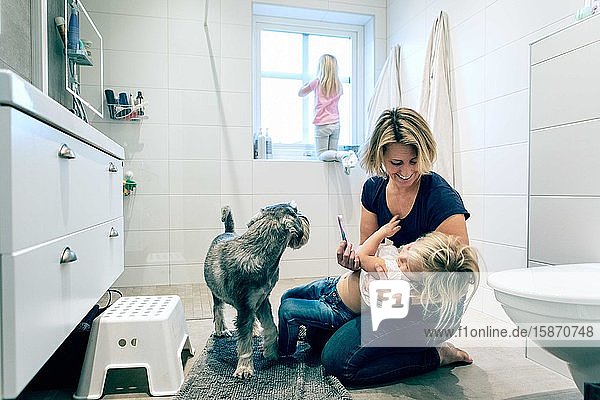 Lächelnde Mutter hält Tochter mit Zahnbürste von Hund im Badezimmer