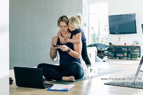 Tochter schaut auf Smartphone  während die Mutter zu Hause auf der Matte sitzt