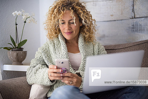 Lächelnde Frau mit Smartphone und Laptop auf dem Sofa