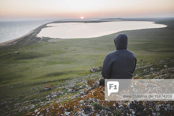 Junger Mann in Kapuzenjacke sitzt bei Sonnenuntergang auf einem Hügel