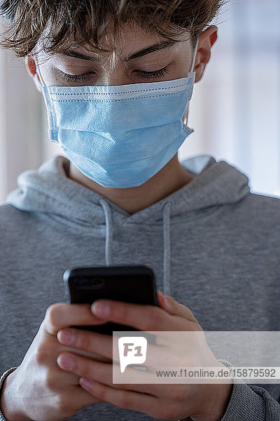 Close up of teenage boy with medical mask using smartphone during coronavirus