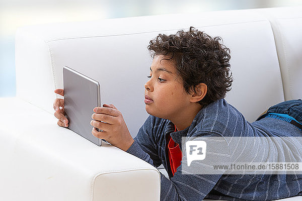 Junger Teenager  der einen Tablet-PC benutzt  während er sich auf seinem Bett entspannt und mit ernster Miene auf dem Bildschirm liest