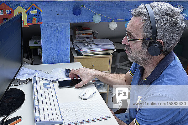 Mann bei Telearbeit zu Hause an seinem Schreibtisch mit Computer  Smartphone und Kopfhörern.