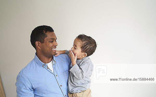 Ein Mann hält seinen Sohn und lächelt ihn an  als der Junge ihm die Hand über den Mund legt.