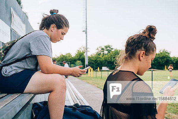 Zwei Frauen sitzen auf Bänken am Rande des Trainingsplatzes und schauen auf ihre Mobiltelefone.
