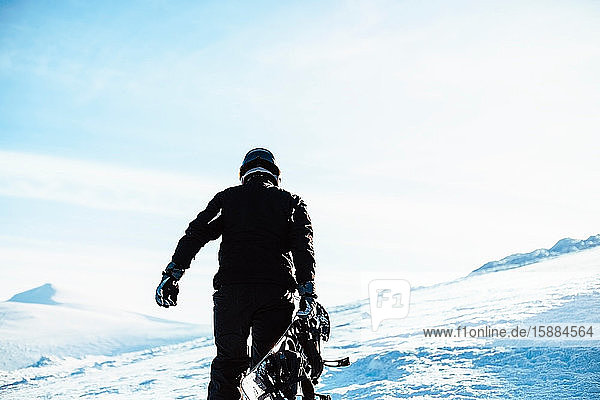 A person wearing a black ski suit and helmet holding a snowboard standing on top of a snowy mountain.