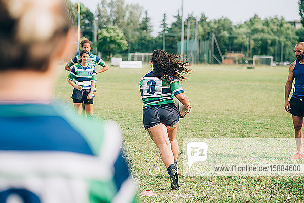 Frauen in blauen  weißen und grünen Rugbyhemden auf einem Trainingsplatz  eine läuft mit einem Rugbyball und der Trainer steht seitlich.