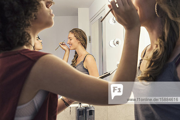 One woman putting make up on another woman with a reflection of them in the mirror.