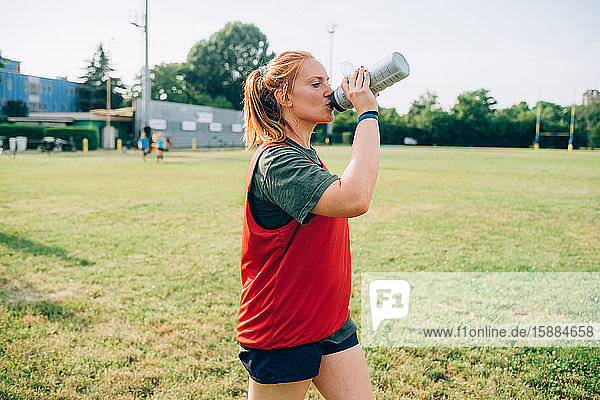 Eine Frau geht über einen Trainingsplatz und trinkt aus einer Wasserflasche.