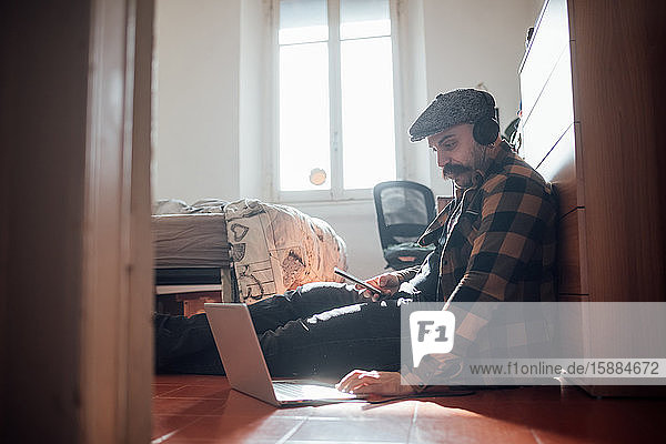Mann mit dickem Schnurrbart und flacher Mütze  der während der Corona-Virus-Krise mit Laptop-Computer auf dem Schlafzimmerboden saß.