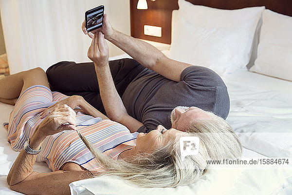 A couple lying on a bed taking a selfie on a mobile phone.