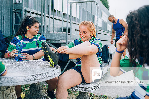 Three women wearing blue  white and green rugby sitting at a table beside a metal grandstand.